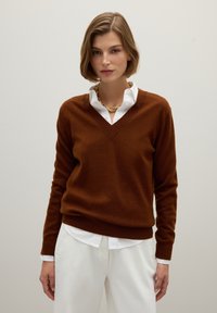 Pull en laine marron avec décolleté en V, manches longues et coupe décontractée, superposé à une chemise blanche à boutons. Chaîne en or visible.