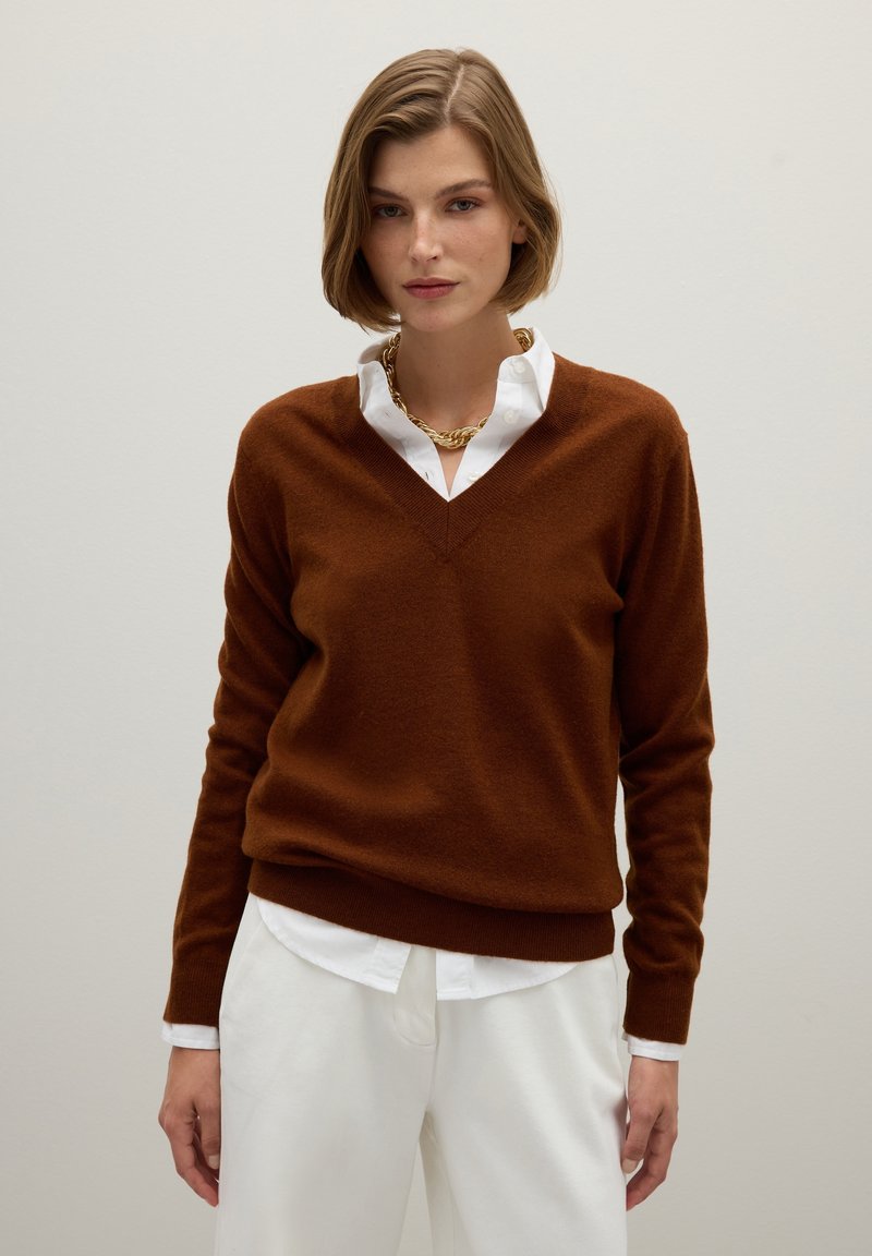 Pull en laine marron avec décolleté en V, manches longues et coupe décontractée, superposé à une chemise blanche à boutons. Chaîne en or visible.