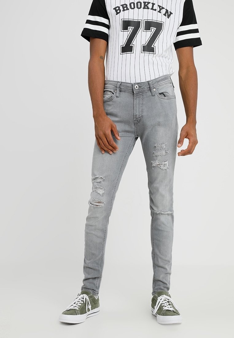 Hombre vistiendo jeans ajustados rasgados grises, camiseta blanca y negra con el texto "Brooklyn 77" y zapatillas verdes con cordones blancos, apoyado contra un fondo liso.