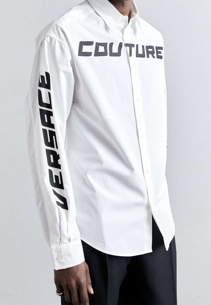 Weißes Hemd mit Knopfleiste, das auf dem Ärmel fett den Schriftzug "VERSACE" und auf der Brust "COUTURE" zeigt; gefertigt aus glattem Stoff.