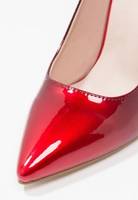 Chaussure à talon haut en cuir verni rouge avec un bout pointu et une finition lisse et brillante, présentant une couture délicate le long du bord.