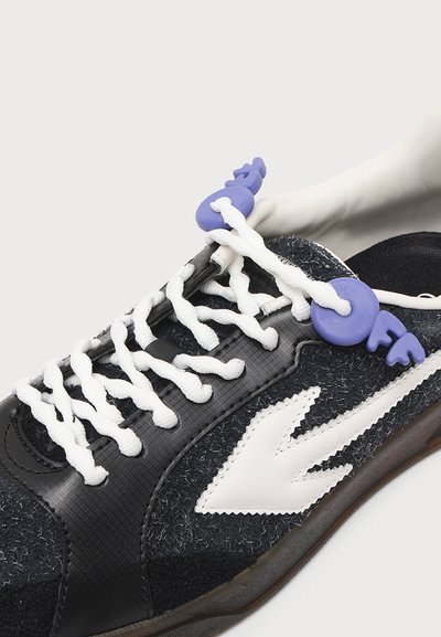 Baskets noires avec des lacets blancs en zigzag et un logo flèche blanc, avec des fermetures de lacets violettes en forme de clés sur fond blanc.