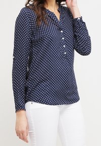 Kvinna som bär en marinblå blus med lång ärm och vitt geometriskt mönster samt vita byxor, som justerar håret med ena handen.