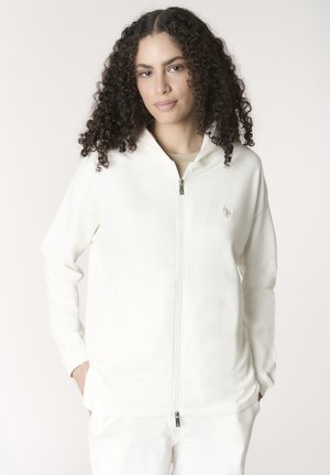 Donna con capelli ricci che indossa una felpa bianca con zip e pantaloni bianchi, in piedi con le mani nelle tasche contro uno sfondo semplice.