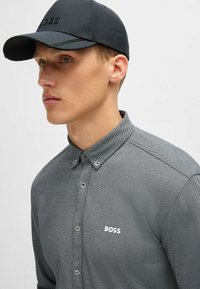 Mörkgrå skjorta med knäppning och en präglad mönstring, med en vit "BOSS"-logotyp och krage. Matchas med en mörk keps.
