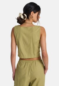 Κοντό τοπ σε ελιά πράσινο με κάθετες ρίγες, στρογγυλεμένη λαιμόκοψη και λεπτομέρειες ραφών. Συνδυασμένο με παντελόνι με ταιριαστές ρίγες.