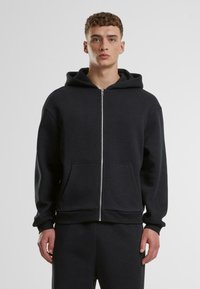 Hoodie noir zippé en tissu doux avec une poche kangourou, une capuche doublée et des poignets côtelés. Porté avec un pantalon de sport assorti.