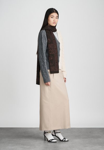 Model draagt een grijze kabelgebreide cardigan over een beige top, gecombineerd met een lange beige rok en zwarte sandaaltjes met bandjes, met een bruine logosc scarf.