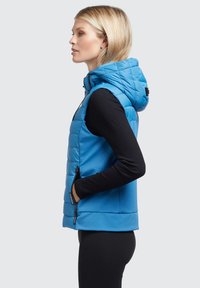Blond kvinna i svarta långärmade tröjor och leggings bär en blå vadderad ärmlös jacka med huva och har händerna i sidofickorna, vänd mot vänster.
