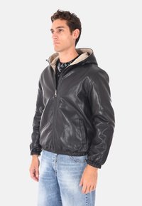 Uomo in giacca di pelle nera con zip e cappuccio, con polsini elasticizzati e jeans azzurri, in piedi davanti a uno sfondo semplice.