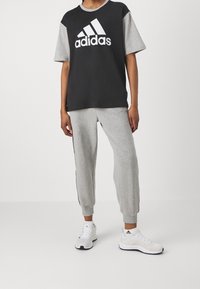 Černé a šedé tričko Adidas s krátkým rukávem a velkým bílým logem, v kombinaci se šedými tepláky s bočními pruhy a bílými sportovními botami.