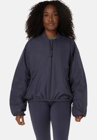 Frau mit lockigem Haar, die eine marineblaue Bomberjacke mit Reißverschluss und passende Leggings trägt, steht vor einem schlichten Hintergrund.