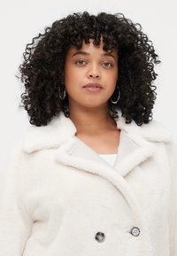 White faux fur coat with a wide collar, double buttons, and a soft texture. Worn over a light top, features hoop earrings.