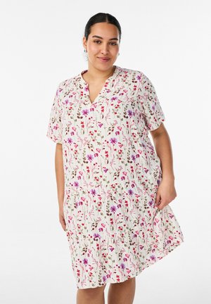 Kvinde iført en hvid knælængde kjole med pink, lilla og rød blomsterprint, står mod en ensfarvet hvid baggrund.