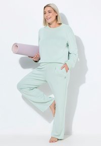 Licht mintgroene sportoutfit bestaande uit een top met lange mouwen en een losse broek. Het model houdt een opgerolde, gestructureerde paarse yogamat vast.