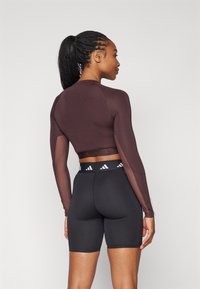 Dugulji smeđi crop top s dugim rukavima i prozirnim rukavima te crne šortsice viskog struka s elastičnim pojasom s logom. Glatki, prilagođeni materijal, atletski dizajn.