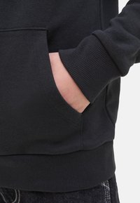 Sweat-shirt noir avec des poignets et un ourlet côtelés, doté d'une poche avant. Le tissu semble lisse et légèrement texturé.