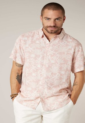 Man met kort haar en baard, gekleed in een lichtroze overhemd met tropische print en korte mouwen, en een witte broek met handen in zijn zakken.