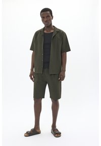 Camisa de manga corta verde oliva con un patrón texturizado, combinada con pantalones cortos a juego. Llevada sobre una camiseta negra, completada con sandalias marrones.