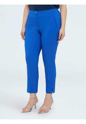 Woman wearing bright blue slim-fit ankle pants and silver high-heeled sandals against a plain background.