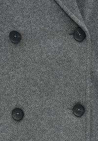 Kaksirivinen harmaa villakangastakki, jossa on pehmeä tekstuuri ja kaksi mustaa nappia edessä. Siistit kauluskäänteet ja hienovaraiset ompeludetaljit.