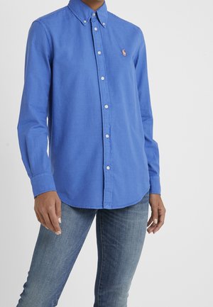 Person wearing a blue long-sleeve button-up shirt with collar and light blue jeans, standing against a plain light background.