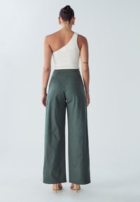 Groene wijde broek van textuurstof, gecombineerd met een witte geribbelde top met één schouder. De schoenen zijn huidskleurig met een spitse neus.