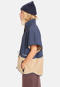 Marineblauwe en beige fleece jack met rits, korte mouwen, hoge kraag, voorzakken en een trekkoord aan de onderkant. Bevat het merklogo aan de onderrand.