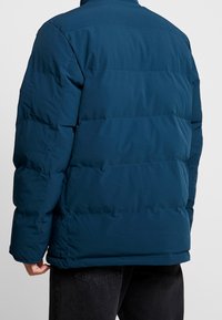 Persona con una chaqueta de invierno acolchada en verde azulado oscuro vista desde atrás, combinada con pantalones negros sobre un fondo liso.