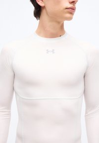 Νεαρός ενήλικας που φοράει στενή λευκή αθλητική μπλούζα με μακριά μανίκια και το λογότυπο της Under Armour στο στήθος, κοιτάζοντας προς τα δεξιά.