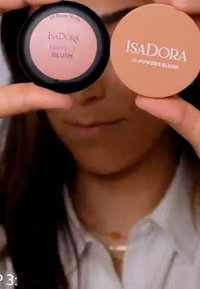 Due compatti di blush IsaDora: un blush in polvere rotondo color pesca con il logo sul coperchio e un pan nero di blush in polvere in una delicata tonalità di rosa.