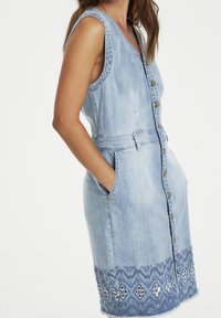 Džínové šaty bez rukávů, světle modré barvy, s předním zapínáním na knoflíky, bočními kapsami a vyšívaným lemem s geometrickými vzory.