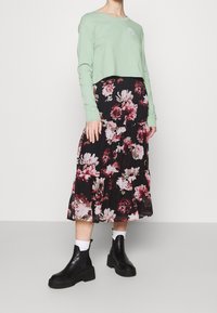 Zelený krátký top s dlouhým rukávem spárovaný s černou midi sukní s květinovým potiskem, která obsahuje růžové a burgundské květiny; oblečeno s černými kotníkovými botami.