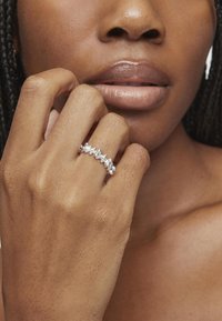 Srebrny pierścionek z wieloma błyszczącymi kamieniami ułożonymi w rzędzie, noszony na dłoni o jasnym odcieniu skóry, ukazujący gładką teksturę skóry.