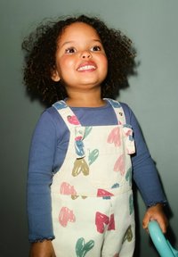 Otrok, oblečen v modro majico z dolgimi rokavi in kremne kombinezone s pisanimi vzorci listov. Kodrasti lasje, nasmeh na obrazu, drži turkizni predmet.