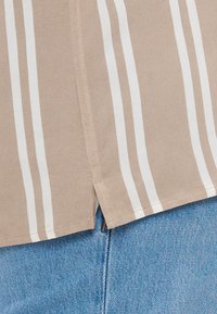 Camisa beige con rayas verticales blancas, con dobladillo recto y abertura lateral, combinada con denim azul claro. Textura de tela suave evidente.