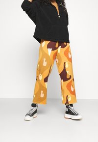 Sudadera de forro polar negra con media cremallera; combinada con pantalones de pierna ancha con estampado en formas abstractas de color naranja, marrón y crema. Zapatillas negras.