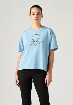 Jonge vrouw draagt een lichtblauw T-shirt met een hoofdtelefoongrafiek en zwarte broek, staand tegen een effen grijze achtergrond.