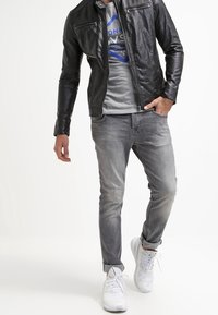 Černá kožená bunda s kapsami na zip, šedé tričko s modrou grafikou, světle šedé džíny a bílé tenisky.