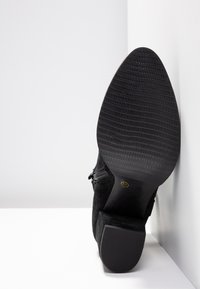 Bottines noires en suede avec une semelle en caoutchouc texturée, dotées d'un talon carré et d'une fermeture éclair sur le côté. La semelle présente un motif pour une meilleure adhérence.