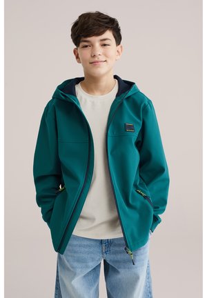 Jugendlicher Junge steht mit Händen in den Taschen, trägt eine türkisfarbene Kapuzenjacke über einem beigen Hemd und hellblauen Jeans, neutraler Hintergrund.