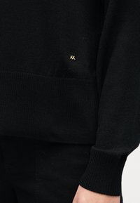 Svart strikket genser med ribbet kant og mansjetter. Har en liten gull "AX"-logo på siden. Teksturen er jevn og litt tykk.