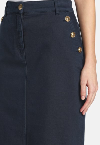 Jupe bleu marine avec des boutons dorés le long de la couture latérale et de la ceinture, portée par une personne dont la main repose à côté.