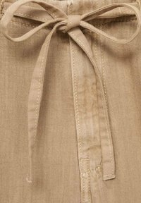 Beige cotton trousers with a fabric tie at the waist. Plain texture with visible stitching details and a smooth finish.