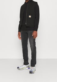 Man wearing black hoodie, black vest with logo patch, dark grey jeans, and white, blue, and yellow running shoes standing on white floor.