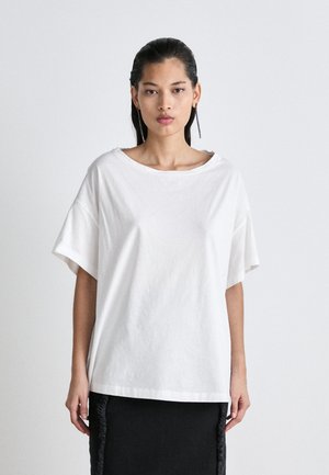 T-shirt bianco oversize in cotone con scollatura ampia e maniche corte. Tessuto morbido. Indossato con una gonna nera texturizzata.
