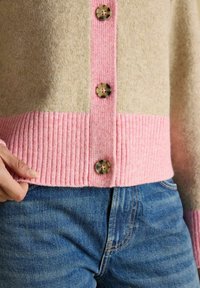 Béžový a ružový pletený kardigan s guličkami v tvare škrupiny nosený s modrými džínami, ruka drží okraj kardiganu na ľavej strane.