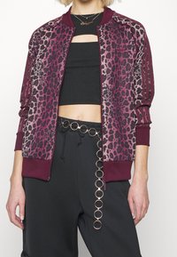 Bomber bunda s leopardím vzorem v fialové a černé barvě, se žebrovanými manžetami a límcem, zapínáním na zip a kontrastními pruhy. Kombinováno s černými kalhotami a řetízkovým páskem.