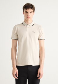 Beige polo ing klasszikus gallérral, fekete díszítéssel a galléron és az ujjakon, bal mellen egy kis fekete logóval.