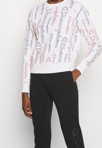 Sweatshirt bianco con il logo "Champion" colorato in rosa e blu scuro, maniche lunghe, polsini a coste e taglio corto. Abbinato a pantaloni neri.